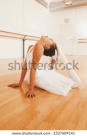 Side view of teenage ballet dancer in tutu and pointe shoes stretching body during lesson in dance hall looking up.