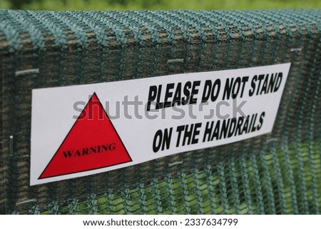 Laminated sign "Please do not stand on the handrails" green netting in background