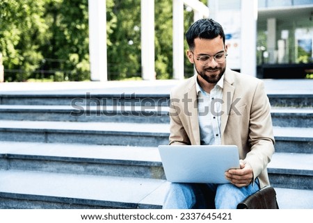 Handsome businessman in glasses working on his laptop computer while resting in city centre. Freelance man typing something in his document. Office worker finishing work outside of office building.