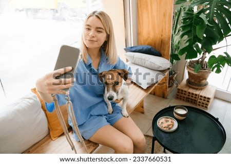 Beautiful young girl taking selfie with her small dog Jack Russell terrier. Comfortable blue clothes. Lunch coffee break in cafe. Influencer blogger live streaming. Horizontal composition photo. 