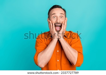 Photo of funky shocked man dressed orange shirt arms cheeks empty space isolated turquoise color background