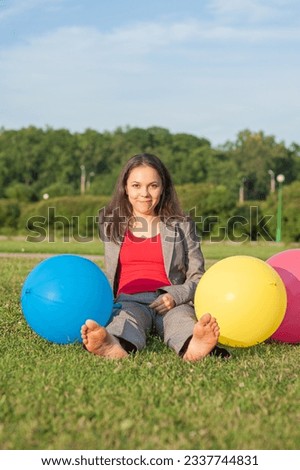 Young beautiful happy barefoot business woman with long hair sitting on the grass with air balloons outdoor in the city park