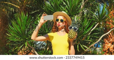 Portrait of beautiful young woman taking selfie with phone and pineapple in sunglasses and blowing her lips sends kiss in summer park on palm tree background