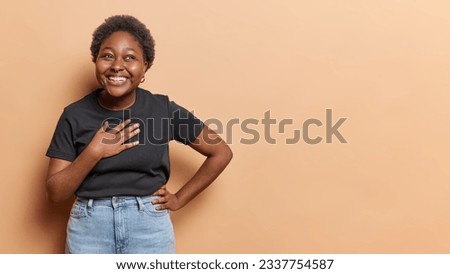 Horizontal shot of cheerful plump woman keeps hand on chest being in good mood laughs happily dressed in black t shirt and jeans isolated over brown background copy space for your advertisement