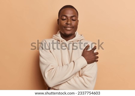 Serene dark skinned man dressed in casual brown sweatshirt embraces himself has eyes closed finding comfort and solace epitomizes inner peace and self acceptance isolated over beige background Royalty-Free Stock Photo #2337754603