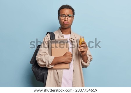 Frustrated disappointed African man holds textbooks spiral notebooks and bottle of orange juice purses lips wears brown shirt carries rucksack isolated over blue background. Studying lifestyle concept Royalty-Free Stock Photo #2337754619