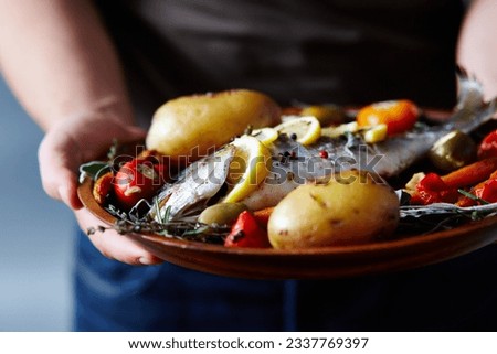 Close-up view of unrecognizable man holding plate with delicious fish baked with potatoes, vegetables, spices and lemon