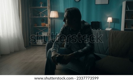 A young man sits in a dark room on the couch with a phone in his hands. The man looks out the window, realizing that it has been a day using the phone, he did not notice how time flew by.