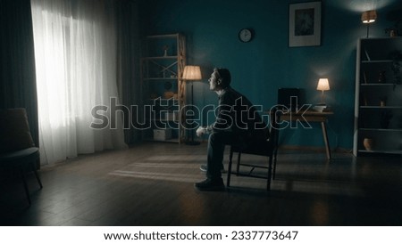 A man sits on a chair in the middle of the room and looks at the light in the window. Side view of a lonely man sitting in a dark room. Hope concept, mental health. Royalty-Free Stock Photo #2337773647