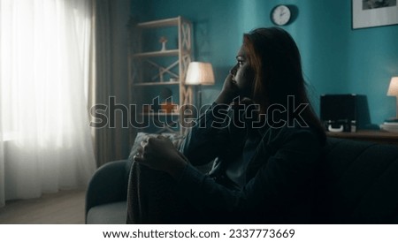 A devastated, distant, lonely young woman sits on a sofa in a dark room close up. A woman desperately thinks about personal problems, experiencing a crisis, looks at the light pouring from the window. Royalty-Free Stock Photo #2337773669