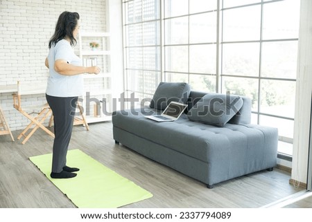 Asian senior lady, elderly woman exercising or working out on Yoga mat while watching fitness videos on her laptop on sofa and workout along VDO online, Senior heathy by exercise at home concept.
