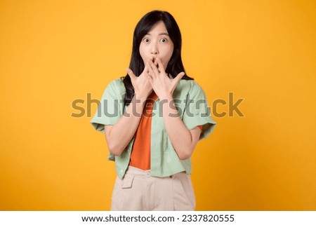 surprise and excitement 30s young Asian woman, wearing green shirt on orange shirt, with a surprised expression and her hand close to her mouth. concept of surprise promotion against yellow backdrop. Royalty-Free Stock Photo #2337820555