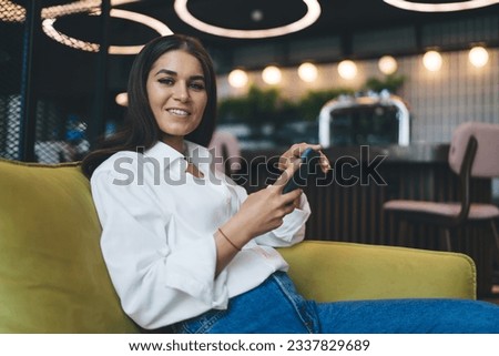Positive young female smiling and looking at camera while relaxing in comfortable sofa against blurred modern cafe with mobile phone in hands