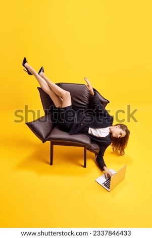 Young business woman lying on armchair in uncomfortable position, working on laptop against bright yellow background. Concept of business, working routine, deadlines, freelance, office, ad Royalty-Free Stock Photo #2337846433