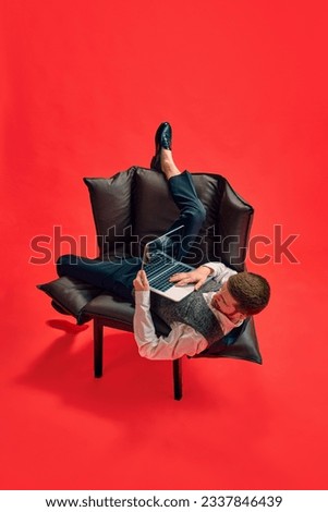 Businessman, employee sitting on armchair in weird, uncomfortable position, working on laptop against red studio background. Concept of business, working routine, deadlines, freelance, office, ad Royalty-Free Stock Photo #2337846439