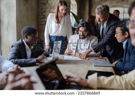 Diverse Business Team Signing Documents - A multicultural group with laptops, a young Middle-Eastern entrepreneur signs papers guided by a senior.