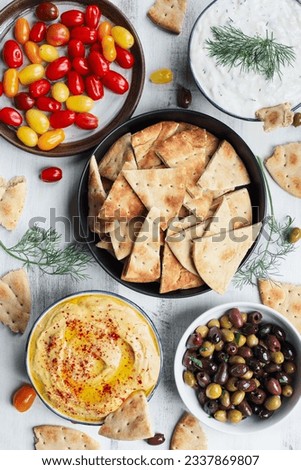 Mezze platter of pita bread surrounded by fresh tomatoes, olives, vegan tzatziki dip, and hummus over a white rustic table. Flatlay. Royalty-Free Stock Photo #2337869807