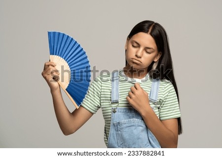 Sweaty overheated teenage girl using paper fan suffer from heat feels sluggish. Displeased tired teen girl in overalls cooling in hot summer weather isolated on studio gray background. Stuffiness. Royalty-Free Stock Photo #2337882841