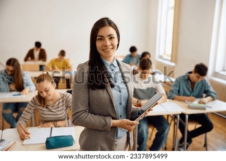 Smiling female teacher with touchpad during a class at high school looking at camera. Her student are learning in the background.