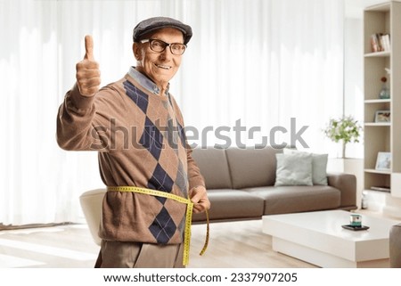 Elderly man measuring waist with a tape inside a living room and gesturing thumbs up