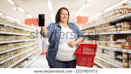 Corpulent woman with a shopping basket showing a smartphone inside a supermarket