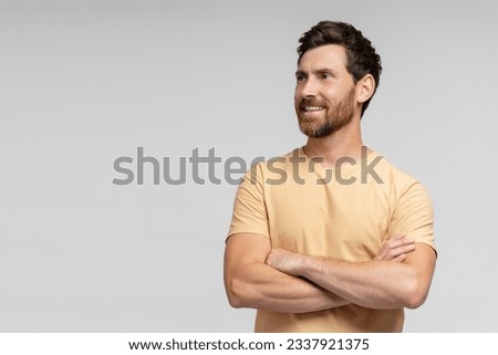Smiling handsome bearded man, hipster wearing blank t shirt looking away and posing arms crossed isolated on gray background. Happy fashion model posing for pictures, studio shot. Mockup 