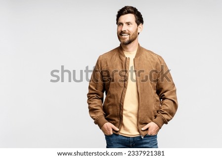 Handsome smiling bearded man wearing brown autumn jacket, stylish jeans isolated on gray background. Portrait of successful middle aged fashion model posing for pictures, studio shot 