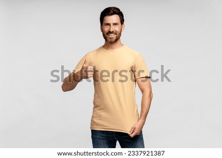 Portrait of smiling handsome bearded man, hipster wearing blank t shirt looking at camera with cool gesture isolated on gray background. Happy fashion model posing for pictures, studio shot. Mockup 