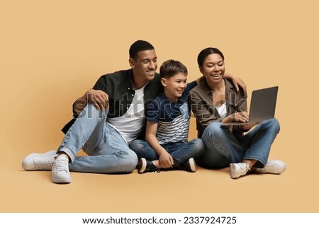 Happy cheerful young black family father, mother abd preteen child son sitting on floor over beige studio background, choosing movie cartoon online, using computer laptop. Family entertainment