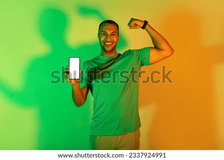 Check this fitness app. Smiling young black man demonstrating smartphone screen and biceps muscles on green and yellow light in studio, looking at camera, advertising workout application. Mockup