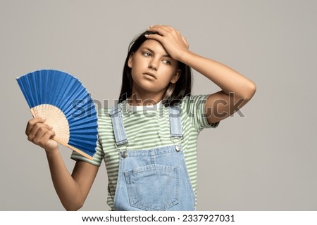 Too hot. Sweaty tired teenage girl touching forehead using paper fan suffer from heat, feels sluggish. Displeased teen girl cooling in hot summer weather, isolated on studio gray wall. Overheating Royalty-Free Stock Photo #2337927031