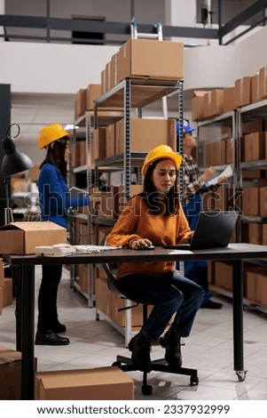 Retail storehouse asian employee managing products catalog on laptop. Postal service young worker wearing yellow protective helmet doing real time inventory tracking on computer Royalty-Free Stock Photo #2337932999