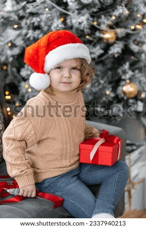 Portrait of a cute little girl in a beautiful knitted sweater ad a red Santa hat near decorated Christmas and new year tree at home. Smiling, cheerful and happy, holding red gift box