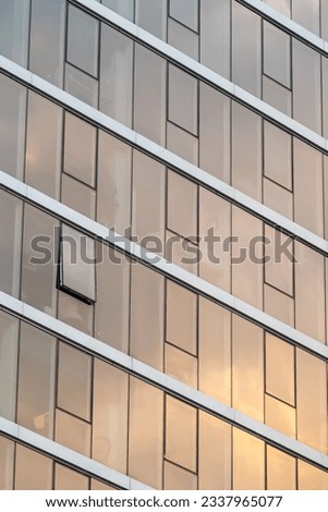 Abstract view of a modern urban apartment building with rows of glass windows reflecting sunset shades of orange, pink and grey. One window open while the rest are closed. Vertical orientation.