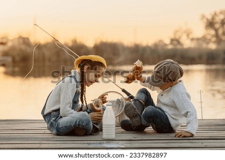 cute blonde boy and girl fishermen in caps and rubber boots eat a bun and drink milk from a glass bottle on a wooden pier, in the morning at dawn, the concept of active local recreation