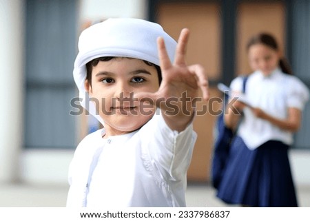 Student wearing Thawb Kandura at school with raised three finger gesture with blurred girl classmate at the background. Arab boy at school premises for educational concept Royalty-Free Stock Photo #2337986837