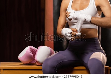 Determined Asian female Muay Thai boxer with muscularity physical readiness body wraps her hand and dons or wearing boxing glove, preparing for intense boxing training in the ring at gym. Impetus Royalty-Free Stock Photo #2337995049