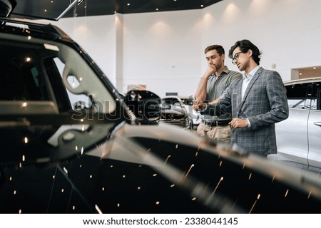 View from hood of car to male dealer in business suit having conversation with pensive young man client at showroom. Doubtful customer guy listening salesman while buying auto at dealership center Royalty-Free Stock Photo #2338044145