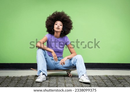 Full body of young African female in Afro hairstyle and casual clothes with makeup sitting on skateboard over terraced floor and looking at camera against green wall Royalty-Free Stock Photo #2338099375