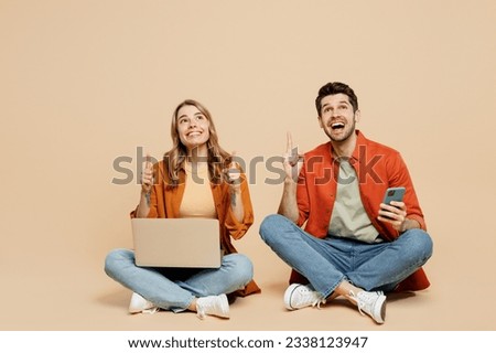 Full body young couple two friend family IT man woman wearing casual clothes together sit hold work on laptop pc computer use mobile cell phone point overhead isolated on pastel plain beige background