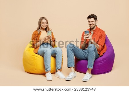 Full body happy young couple two friends family man woman wear casual clothes together sit in bag chair hold in hand use mobile cell phone isolated on pastel plain light beige color background studio