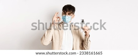 Health, covid and quarantine concept. Portrait of natural guy in glasses and face mask showing bottle of good hand sanitizer, make okay sign, recommending product, white background.