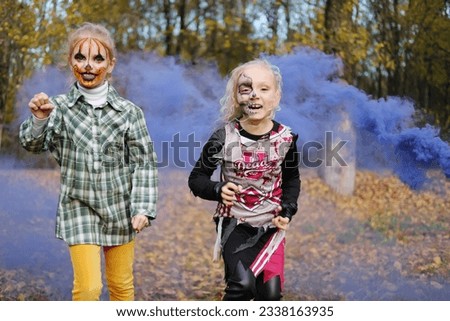Two children girls girlfriends in Halloween costumes and with makeup make frightening grimaces against the background of autumn leaves and purple smoke. Horizontal photo