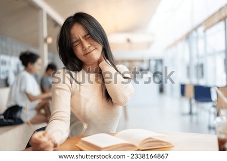 A tired and unhappy young Asian female college student suffering from a sore neck after a long reading session sitting in a coffee shop or library. Health care and education concept Royalty-Free Stock Photo #2338164867