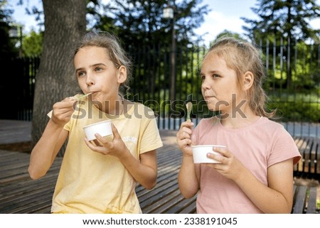portrait of two cute blonde girls, they eat cold ice cream from paper cups. A walk in the park, summer holidays and fun