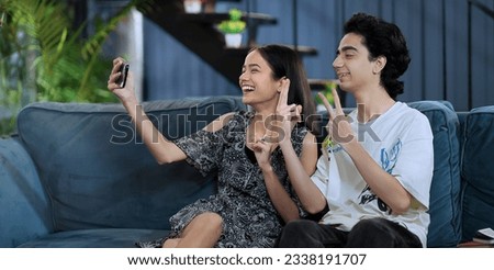 Smiling girlfriend sitting on sofa taking funny faces selfie with teen boy using mobile phone at indoor home. Happy Indian teenager girl making self photos record vlog on smartphone enjoy fun together