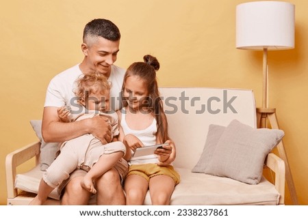 Smiling father with child daughters wearing casual clothing sitting on sofa against beige background family using mobile phone watching cartoons together.