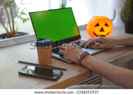 the hands of a young programmer work on the keyboard of a laptop with a green screen on a wooden table near the window in a cafe against the background of a glowing pumpkin with a smiling  face.
