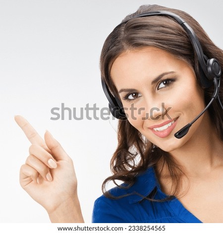Portrait of happy smiling beautiful young support phone operator showing blank copyspace area for slogan or text, posing at studio against grey background Royalty-Free Stock Photo #2338254565