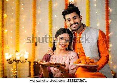 Happy smiling indian couple in traditional ethnic wear by holding sweets at home - concept of diwali festive celebration, greeting and family relationship Royalty-Free Stock Photo #2338273671